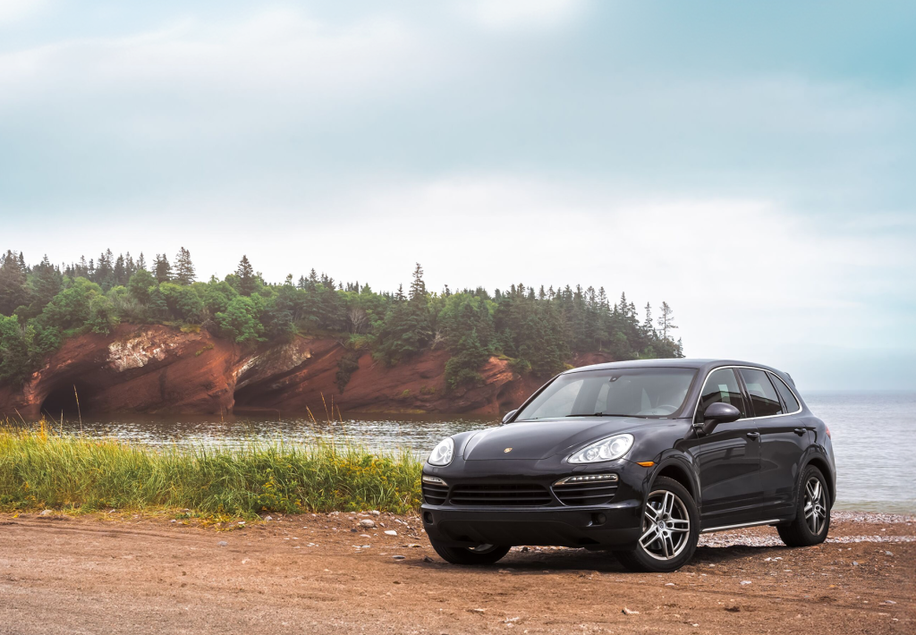 A black SUV is parked on a shoreline, with a tall, rocky peninsula behind it.