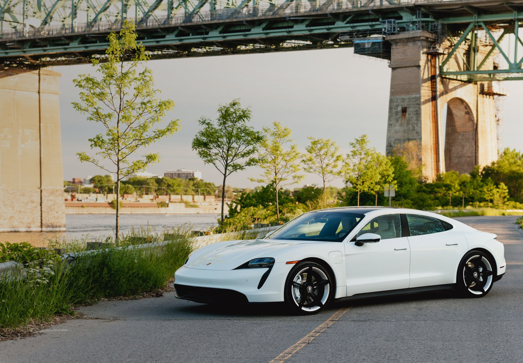 A white sports car is parked on a sunny, tree-lined road below a bridge.