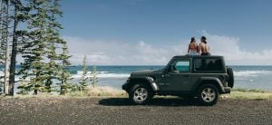 A man and woman sit on top of their green SUV overlooking the ocean.