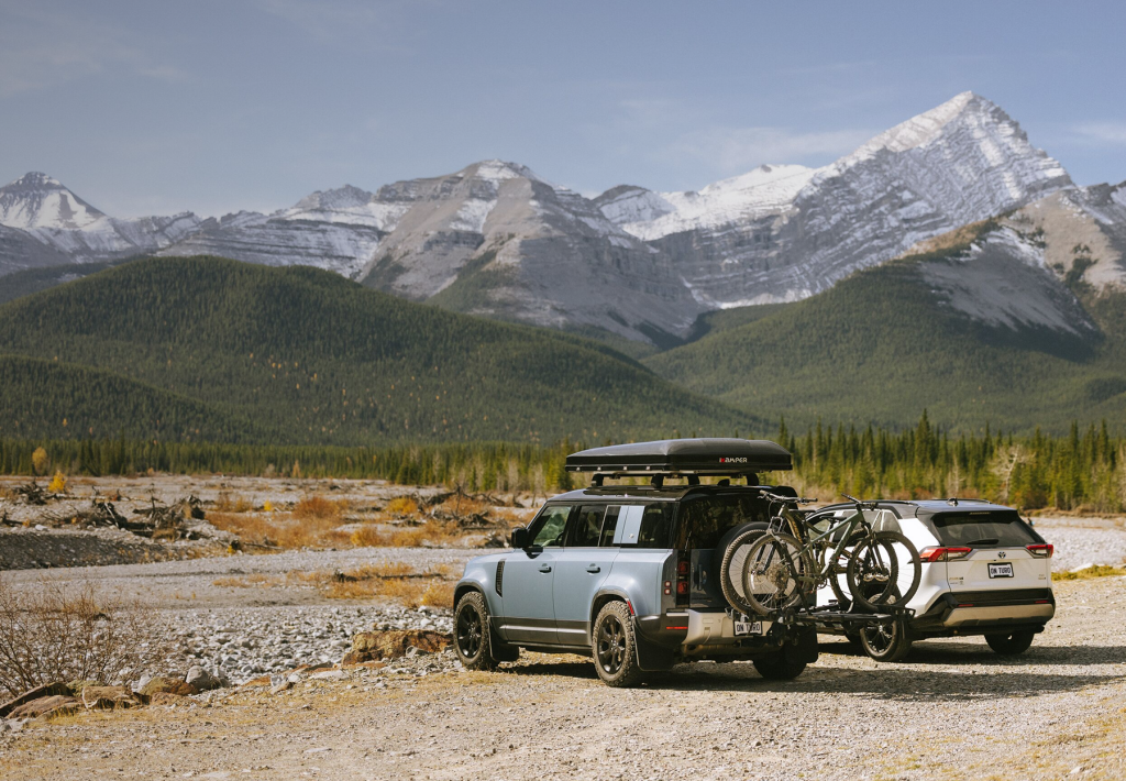 Two SUVs, one with a rooftop carrier and bikes on the trunk, are parked in front of a mountain.