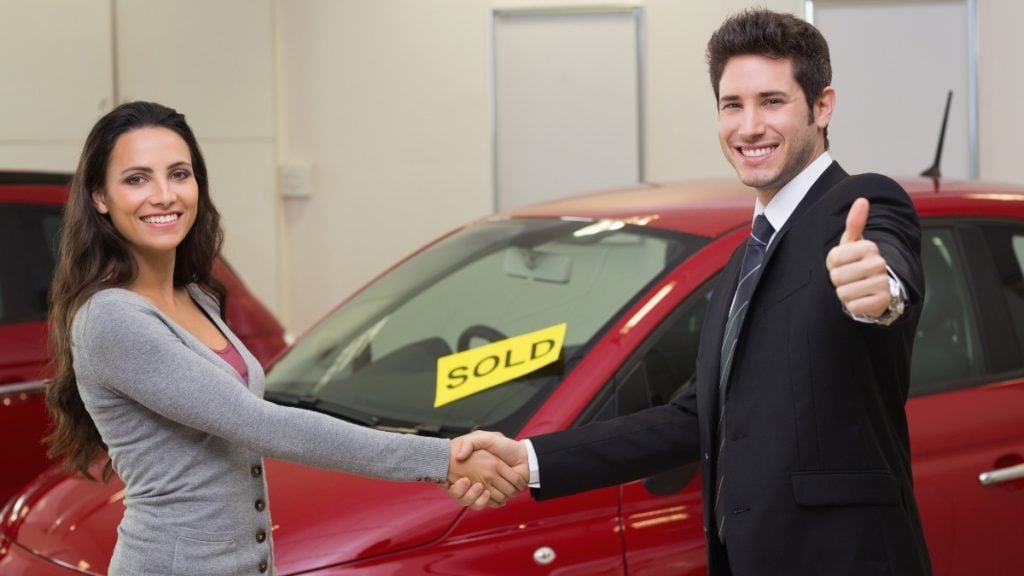 Buyer and dealer shaking hands in front of a sold car at a dealership