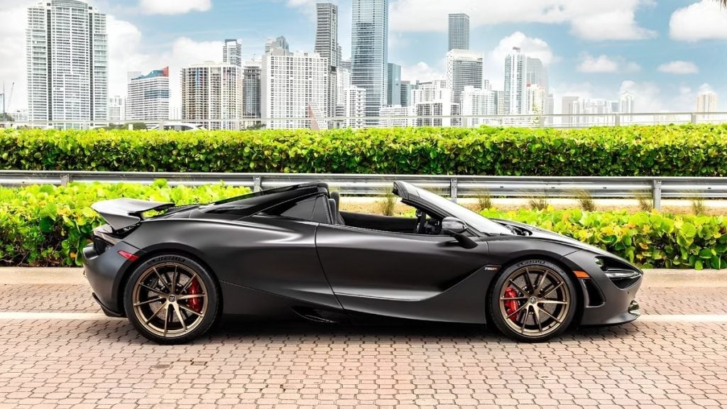 Side profile of a matte black McLaren 720S Spider with roof down, showing its sculpted carbon fiber body against the Miami skyline